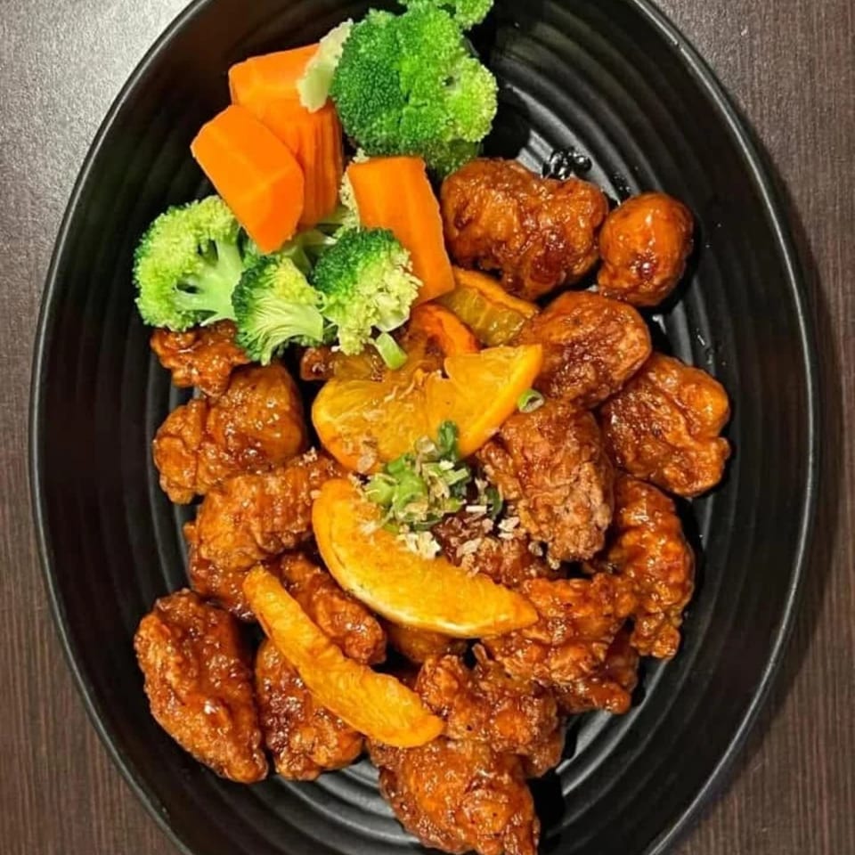 Orange chicken with broccoli, carrots, and orange slices on a black plate.